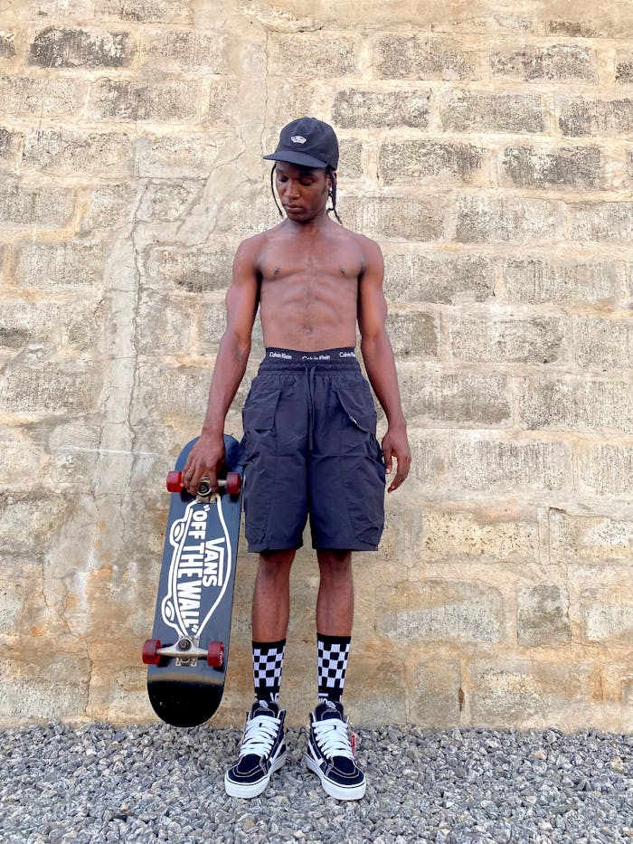 African adult male skater standing shirtless against brick wall holding a skateboard.