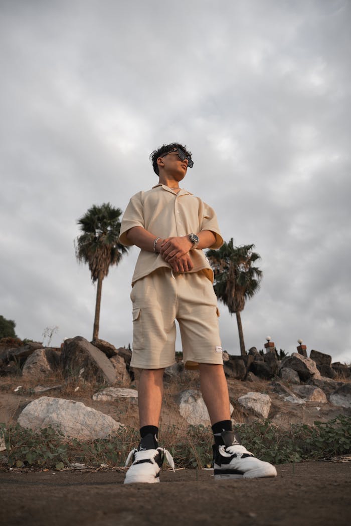 Young man in casual attire confidently stands outdoors against a backdrop of palm trees and rocks.