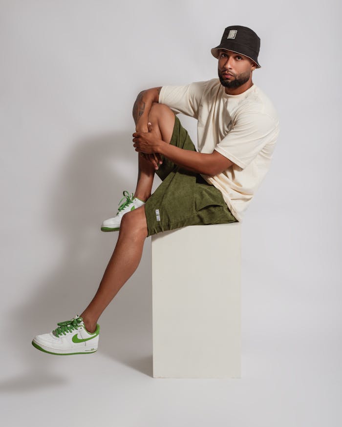 Man sitting on a white box, wearing casual fashion. Stylish outfit with sneakers and bucket hat.